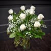 Tall arrangement of white roses and small white filler flowers in a clear glass vase.