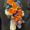 Tall cross-shaped spray of white carnations with orange lilies, roses, and blue flowers on a stand