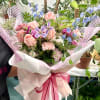Hand-tied bouquet of peach roses and pastel mixed flowers wrapped in white and pink paper with a magenta ribbon