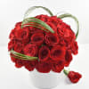Compact arrangement of red roses in a white vase with looping green blades of grass.