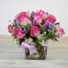 Compact arrangement of hot pink roses with purple blooms in a square glass vase with ribbon.