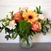 Peach roses, pale carnations, a yellow gerbera daisy, and a green pompom bloom in a clear glass jar.