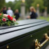 Small spray of pink and red roses on a dark casket at an outdoor funeral.