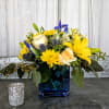 Compact arrangement of yellow lilies, cream roses, and blue accents in a blue glass cube vase.