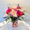 Pink roses and pale gerbera daisies arranged with mixed blooms in a pink glass vase with ribbon.