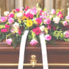 Bright pink and cream rose casket spray with white ribbon on a wooden casket.