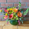 Low arrangement of orange roses, pink calla lilies, and green orchids in a round green glass vase