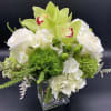 Compact arrangement of white roses, hydrangeas, and green orchids in a clear glass cube vase.