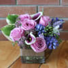 Short arrangement of pink roses, purple calla lilies, and blue flowers in a square glass vase