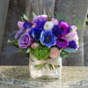 Compact arrangement of purple roses, blue and fuchsia anemones, and peach blooms in a square glass vase.