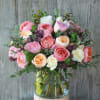 Arrangement of pink, peach, and white roses with purple accents in a clear glass vase