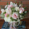 Low pink and white rose arrangement in a clear glass cube vase with ribbon, set on books.