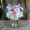 Mason jar arrangement with pink and peach roses, white orchids, hydrangeas, and pink lilies.