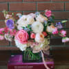 Low pastel arrangement of pink roses and mixed blooms in a square glass vase with ribbon.