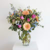 Mixed bouquet of peach gerberas, purple tulips, and blue flowers in a clear glass vase.