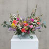 Low, wide arrangement of bright mixed flowers in a ribbed metal container on a white pedestal