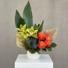 Modern arrangement with green orchids, orange roses, palm fans and large green leaves in a white vase.