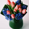 Compact arrangement of pink tulips and blue hyacinths in a glass cylinder vase wrapped with green leaves