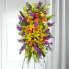 Tall standing spray with yellow lilies, orange roses, and purple and white flowers on a metal easel.