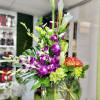 Tall mixed arrangement with white calla lilies, purple orchids, and orange blooms in a round glass vase