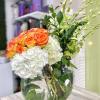 Round glass vase with orange roses, white hydrangeas, and tall green orchid stems