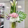 Tall arrangement of white calla lilies, pink roses, orchids, and hydrangeas in a clear cylinder vase.