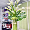 Tall arrangement of white calla lilies with large glossy leaves in a clear glass vase.