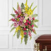 Standing spray of pink lilies, roses, and yellow flowers on a white easel beside a wooden casket.