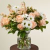 Peach roses, white daisies, and pastel blooms arranged in a clear glass vase.