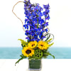 Tall blue delphinium and yellow sunflowers arranged in a clear rectangular glass vase