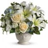 Ivory lilies, cream roses, and white carnations arranged with blue hydrangea in a white ceramic urn vase