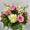 Compact arrangement of pink roses, hydrangea, and mixed pastel blooms in a clear glass vase