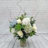 Tall glass vase arrangement with white roses, blue and white hydrangeas, green blooms, and curly willow branches.