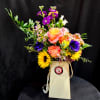 Bright mixed bouquet with roses, sunflowers and purple blooms in a tall cream gift bag-style vase.