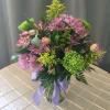 Mixed bouquet of lavender daisies, green mums, and pink blooms in a clear vase with a lavender ribbon