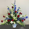 Tall red, white, and blue arrangement with roses, lilies, and delphinium in a white urn vase