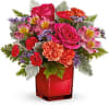 Compact arrangement of pink roses, orange carnations, and mixed blooms in a red cube vase