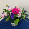 Low glass vase of pink gerbera daisy, roses, and mixed blooms with purple and yellow accents.