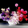 Asymmetrical bowl arrangement of pink, purple and red roses, dahlias and orchids in a white vessel.
