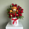 Round box of red roses with small white blooms and a teddy bear nestled in the center.