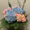 Pastel blue and pink hydrangeas with pink roses and eucalyptus in a clear glass vase with a fern collar