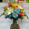 Bouquet of blue hydrangeas, white lilies, and orange roses in a glass vase