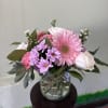 Pink gerbera daisies, roses, and lavender mums in a clear glass vase on a small stand