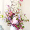 Tall pastel pink and purple flower arrangement in a white ceramic face vase on a rustic crate