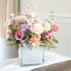 Low pastel arrangement of roses and lisianthus in a frosted glass cube vase on a draped table