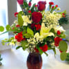 Red roses, white lilies, and red carnations arranged with filler blooms in a red glass vase.