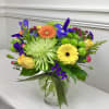 Bright mixed bouquet with yellow roses, gerbera daisies, green spider mums, and purple irises in a glass vase.