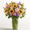 Bouquet of sunflowers, pink roses, and purple flowers in a clear glass vase