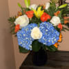 Bouquet of blue hydrangeas, white roses, orange roses, and yellow lilies in a dark vase