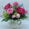 Pink roses, gerbera daisies, green hydrangea, and carnations in a white daisy pitcher with a blue butterfly pick.
